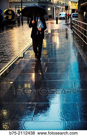 Stock Photo - Unrecognizable man walking in the rain with an umbrella and a folder under his arm. London, England, UK, Europe.. Fotosearch