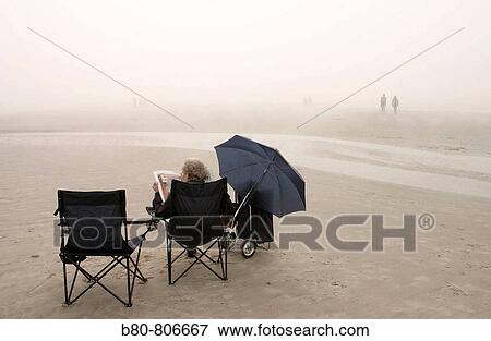 Stock Photo - Weather, fog, expecting the sun, woman, newspaper, chairs, trolley, umbrella, St. Peter-Ording, Schleswig-Holstein, Germany. Fotosearch