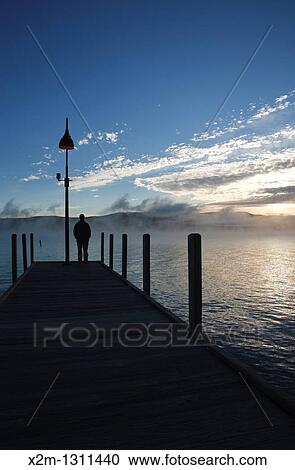 Stock Image - Wellington State Park - The silhouette of a man on dock watching sunrise over Newfound Lake in Bristol, New Hampshire USA. Fotosearch