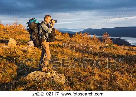 Stock Photo - White man withbackpack taking photo standing on mountain in autumn season in Sweden, Swedish lapland.. Fotosearch