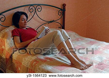 Stock Image - Woman 41 reading a book on her bed, Auvergne, France. Fotosearch