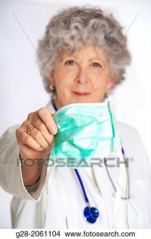 Woman doctor, in a white overall, senior woman with grey hair, and stethoscope around her neck, facing and looking at patient, holding green mask in front of her, in hospital or clinic View Large Photo Image Picture - Woman doctor, in a white overall, senior woman with grey hair, and stethoscope around her neck, facing and looking at patient, holding green mask in front of her, in hospital or clinic. Fotosearch