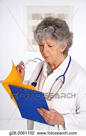 Woman doctor, in a white overall, senior woman with grey hair, red glasses in hand, and stethoscope around her neck, holding and looking at medical files in her hand, in hospital or clinic View Large Photo Image Stock Image - Woman doctor, in a white overall, senior woman with grey hair, red glasses in hand, and stethoscope around her neck, holding and looking at medical files in her hand, in hospital or clinic. Fotosearch