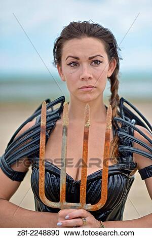 Stock Image - Woman dressed up with a futuristic space look costume holding a rusty trident, creating a modern interpretation of Salacia, Neptune's queen, an ocean Roman deity.. Fotosearch