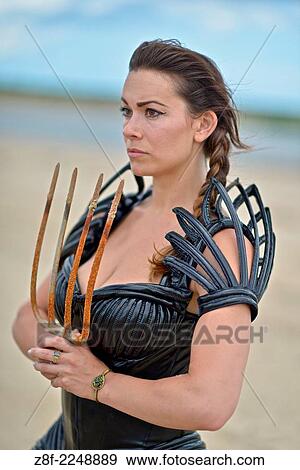 Stock Photo - Woman dressed up with a futuristic space look costume holding a rusty trident, creating a modern interpretation of Salacia, Neptune's queen, an ocean Roman deity.. Fotosearch
