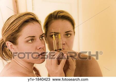 Stock Image - Woman holding a syringe, looking in a mirror. Fotosearch