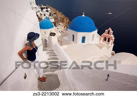 Stock Image - Woman in front of a blue domed church in Oia town with a ferry at the background, Santorini, Cyclades Islands, Greek Islands, Greece, Europe.. Fotosearch