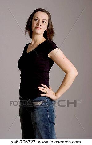 Woman in her early thirties stands confidently, hands on hips, wearing black t-shirt and blue jeans, looking at camera with slight smile. Studio lighting, grey background. View Large Photo Image Stock Photo - Woman in her early thirties stands confidently, hands on hips, wearing black t-shirt and blue jeans, looking at camera with slight smile. Studio lighting, grey background.. Fotosearch