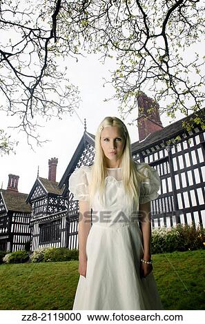 Stock Image - Woman in vintage dress standing outside a tudor house.. Fotosearch