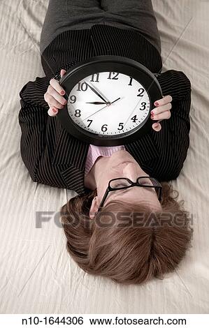 Stock Photograph - Woman lying on her bed, holding a wall clock. Fotosearch