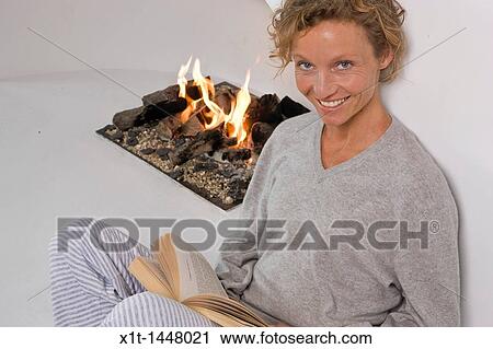 Stock Image - Woman sitting by fireplace, reading. Fotosearch