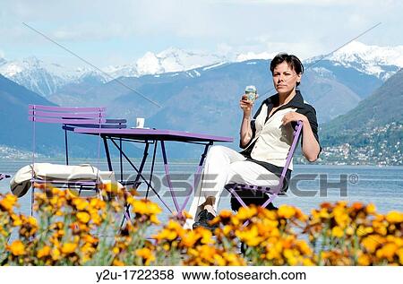 Stock Photo - Woman sitting down in a patio and drinking a glass of champagne close to a lake front with snow-capped mountains. Fotosearch