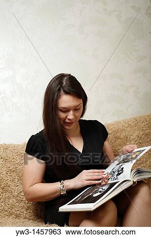 Stock Image - Woman sitting on sofa, looking at book. Fotosearch