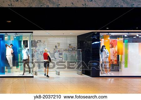 Stock Image - Woman standing at closed shop gate. Mannequins, dummy on store show-window. Retail shop entrance or exit. Facade of shop with display. Exterior.. Fotosearch