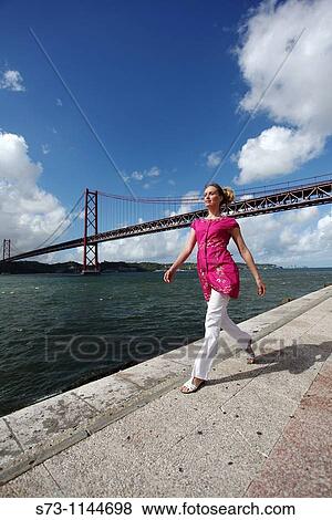 Stock Photo - Woman walking near the 25th April Bridge over the River Tagus, Lisbon, Portugal. Fotosearch