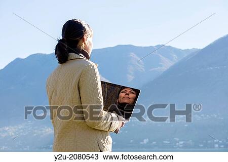 Stock Image - Woman with a computer close to an alpine lake maggiore with mountains and blue sky in ascona ticino switzerland.. Fotosearch