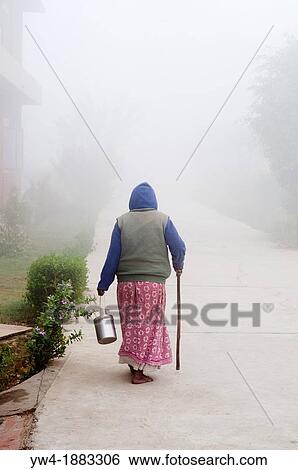 Stock Photograph - Woman with a walking stick on a misty path, Ma Dham Ashram for widows, Vrindavan, Uttar Pradesh, India. Fotosearch