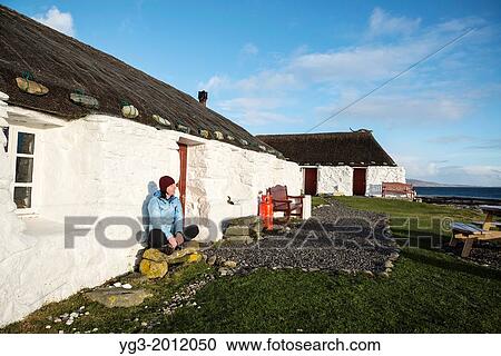 Stock Image - Worman sits infront of thatched roof traditional blackhouse now used as youth hostel, Berneray, Outer Hebrides, Scotland.. Fotosearch