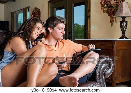 Picture - Young adult couple relaxing in the sofa of their living room, drinking wine and surfing the internet on ipad like tablet computer. Fotosearch