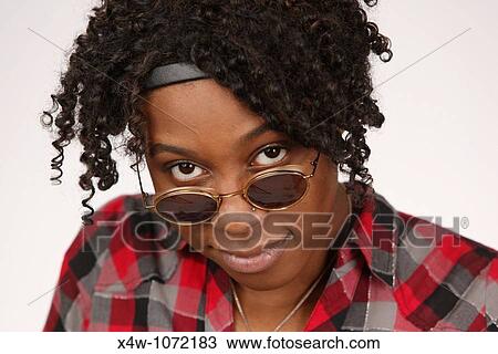 Stock Image - Young African-American woman looking over sunglasses posing for portrait. Fotosearch