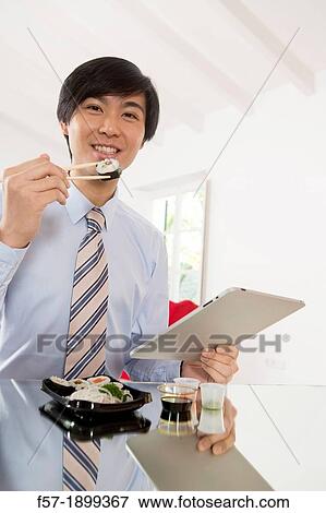 Young Asian businessman eating sushi while communicating on his Tablet computer View Large Photo Image Stock Photo - Young Asian businessman eating sushi while communicating on his Tablet computer. Fotosearch