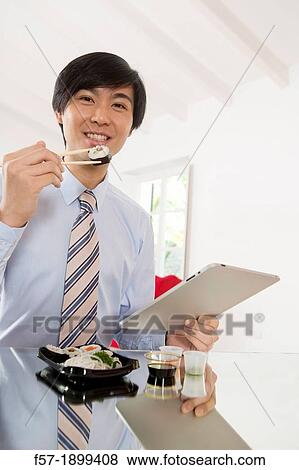 Young Asian businessman eating sushi while communicating on his Tablet computer View Large Photo Image Stock Photo - Young Asian businessman eating sushi while communicating on his Tablet computer. Fotosearch