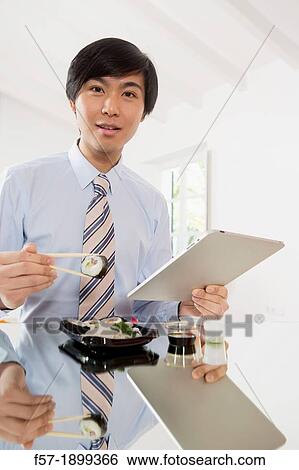Young Asian businessman eating sushi while communicating on his Tablet computer View Large Photo Image Stock Photograph - Young Asian businessman eating sushi while communicating on his Tablet computer. Fotosearch