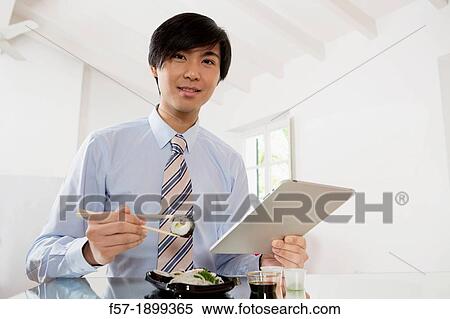 Young Asian businessman eating sushi while communicating on his Tablet computer View Large Photo Image Stock Photography - Young Asian businessman eating sushi while communicating on his Tablet computer. Fotosearch