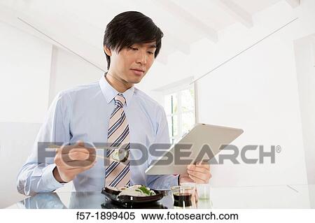 Young Asian businessman eating sushi while communicating on his Tablet computer View Large Photo Image Stock Photography - Young Asian businessman eating sushi while communicating on his Tablet computer. Fotosearch
