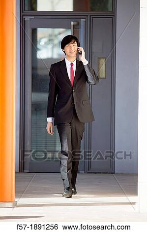 Young Asian businessman walking and talking on his smartphone outside an office building View Large Photo Image Stock Photograph - Young Asian businessman walking and talking on his smartphone outside an office building. Fotosearch