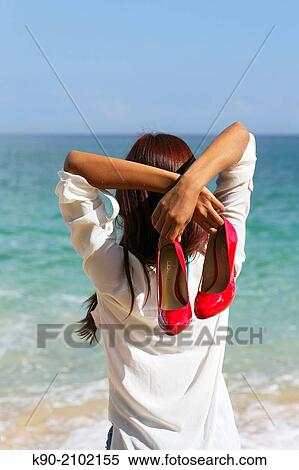 Stock Photography - Young asian woman seen from behind, standing at the beach and holding her red high heals behind her back, Bali, Indonesia, Southeast Asia.. Fotosearch