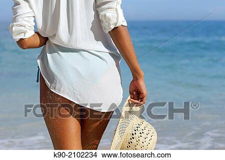 Picture - Young asian woman seen from behind with sand on her legs, holding a hat and standing at the beach, Bali, Indonesia, Southeast Asia.. Fotosearch