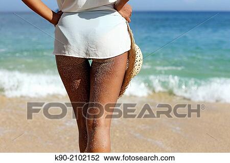 Stock Image - Young asian woman seen from behind with sand on her legs, holding a hat and standing at the beach, Bali, Indonesia, Southeast Asia.. Fotosearch