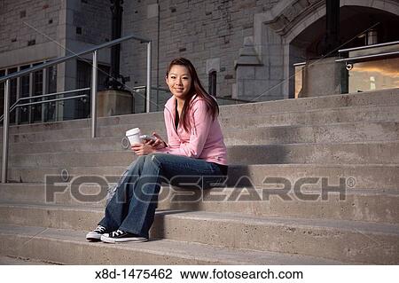 Young Asian woman texting on her smart phone while sitting on outdoor steps with a cup of coffee. View Large Photo Image Stock Image - Young Asian woman texting on her smart phone while sitting on outdoor steps with a cup of coffee.. Fotosearch