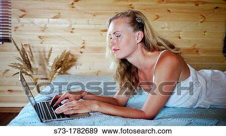 Stock Photo - Young beautiful happy woman lying on the bed with laptop. Fotosearch