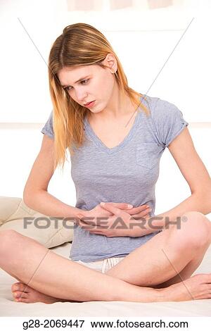 Young beautiful woman sitting on a bed and holding her stomach for pain View Large Photo Image Stock Photo - Young beautiful woman sitting on a bed and holding her stomach for pain. Fotosearch