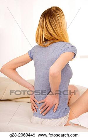 Stock Photography - Young beautiful woman sitting on a bed and suffering from back ache. Fotosearch
