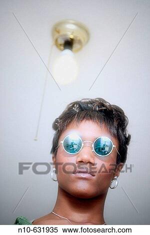 Stock Photography - Young black woman, wearing sunglasses, standing underneath a light fixture.. Fotosearch