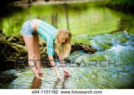 Stock Image - Young blond woman, cut off shorts by a small river in a country outdoor setting. Fotosearch