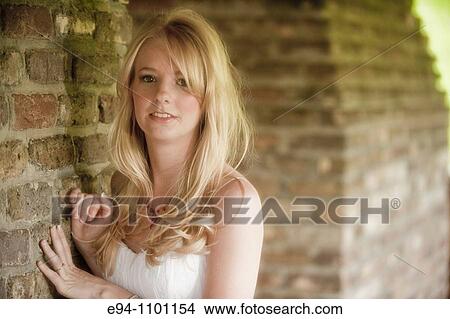 Young blond woman in a white dress smiling camera leaning against a brick wall. View Large Photo Image Picture - Young blond woman in a white dress smiling camera leaning against a brick wall.. Fotosearch