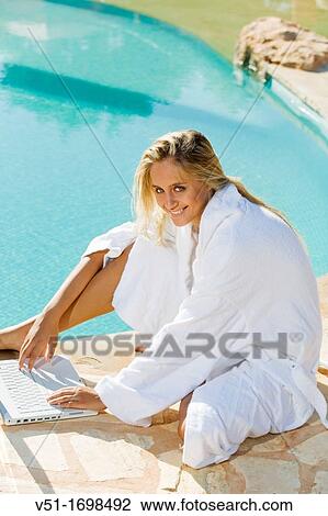 Stock Image - Young blonde woman at pool using laptop and smiling at camera. Fotosearch