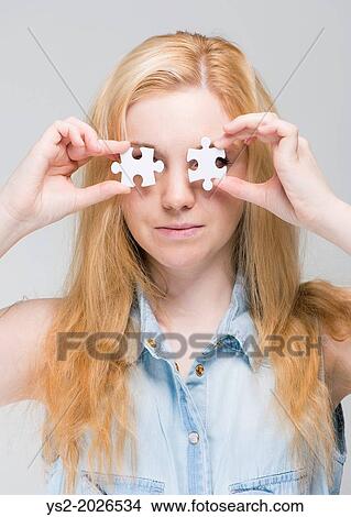 Young blonde woman holding two white puzzle pieces in front of her eyes. View Large Photo Image Picture - Young blonde woman holding two white puzzle pieces in front of her eyes.. Fotosearch