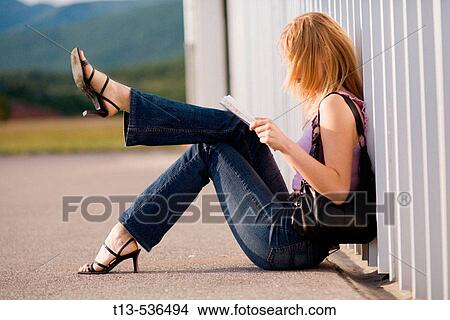 Picture - Young blonde woman is reading a road map while sitting by the wall with one leg lifted hight into air. Fotosearch
