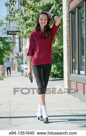 Stock Image - Young brunette woman in a red sweater and short black pants walking on city sidewalk smiling at the camera. Fotosearch