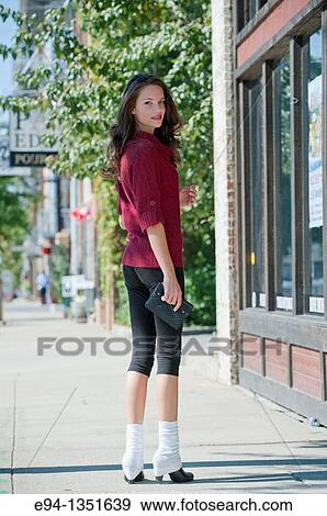 Young brunette woman in a red sweater and short black pants walking on city sidewalk looking over her shoulder smiling at the camera View Large Photo Image Stock Photo - Young brunette woman in a red sweater and short black pants walking on city sidewalk looking over her shoulder smiling at the camera. Fotosearch