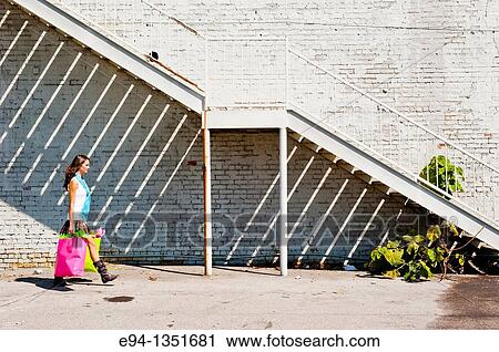 Young brunette woman in a summer dress carrying shopping bags on a sidewalk in an urban setting View Large Photo Image Stock Image - Young brunette woman in a summer dress carrying shopping bags on a sidewalk in an urban setting. Fotosearch
