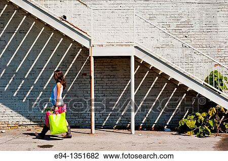 Stock Image - Young brunette woman in a summer dress carrying shopping bags on a sidewalk in an urban setting. Fotosearch
