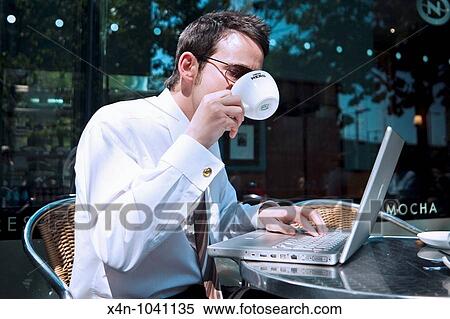 Stock Photography - Young businessman drinks cup of coffee whilst looking at financial data on his laptop outside coffee shop in the City of London, UK. Fotosearch