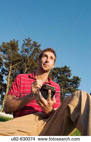 Young Caucasian man sitting on grass holding a handheld PDA while looking out into the distance, San Francisco, California, USA. View Large Photo Image Picture - Young Caucasian man sitting on grass holding a handheld PDA while looking out into the distance, San Francisco, California, USA.. Fotosearch