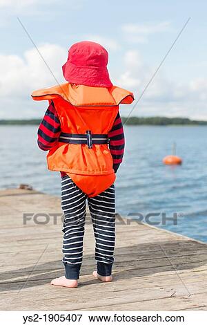 Young child wearing orange life jacket and red hat watching the sea from a wooden jetty View Large Photo Image Stock Photo - Young child wearing orange life jacket and red hat watching the sea from a wooden jetty. Fotosearch